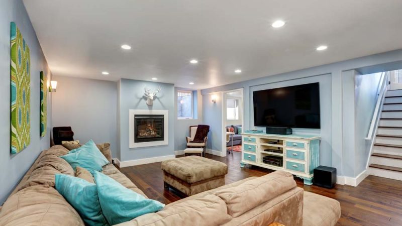 Pastel blue walls in basement living room interior. Large corner sofa with blue pillows and ottoman. Vintage white and blue TV cabinet.